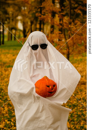Woman in white sheet holding jack o'lantern against autumn forest background. Halloween costume. Vertical photo.  123335570