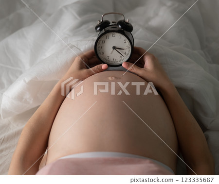 A pregnant woman lies in bed and holds an alarm clock. Close up of the belly. 123335687
