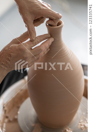 A potter works with a tool on a potter's wheel leveling the surface of a vase. Close-up of a man's hands.  123335741