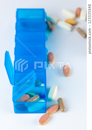 closeup white background two bright blue pill boxes with couple of open lids for taking pills, health care, partial filling, different shapes colors of pills and vitamins pills lie next to the table closeup white background two bright blue pill boxes with couple of open lids for taking pills, health care, partial filling, different shapes colors of pills and vitamins pills lie next to the table 123335848
