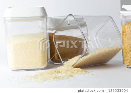 close-up white there are three transparent containers of different sizes with white lids filled with different cereals with rice and brown sugar minimalism spilled rice on the table from a container close-up white there are three transparent containers of different sizes with white lids filled with different cereals with rice and brown sugar minimalism spilled rice on the table from a container 123335904