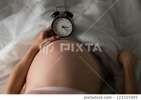 A pregnant woman lies in bed and holds an alarm clock. Close up of the belly. 123335905