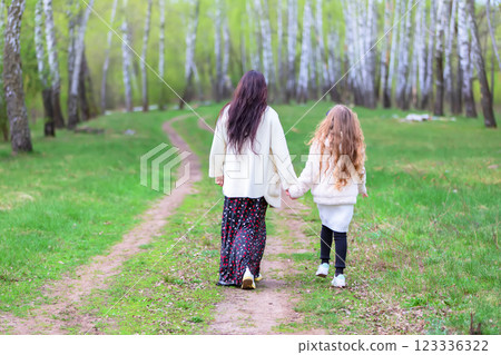 Mom and daughter are walking through a birch grove with their backs to the camera. 123336322