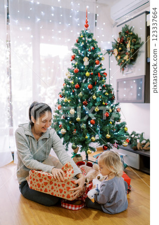 Little girl is watching her mother unpacking gifts while sitting on the floor near the Christmas tree 123337664