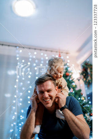 Laughing dad with a little girl on his shoulders leaned forward sitting near the Christmas tree 123337728
