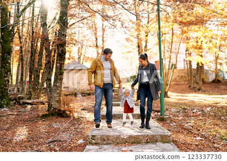 Parents lead their little daughter by the hands along a paved path in the autumn forest, lifting her above the steps Parents lead their little daughter by the hands along a paved path in the autumn forest, lifting her above the steps 123337730