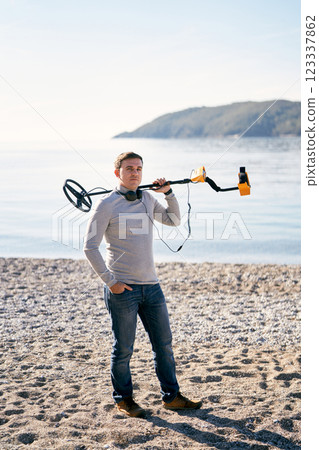 Man with headphones around his neck stands on the seashore with a metal detector on his shoulder 123337862