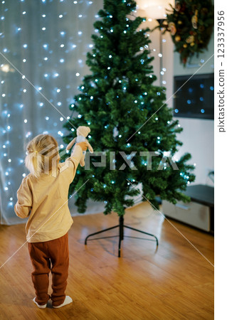 Little girl shows a teddy bear a Christmas tree decorated with sparkling garlands. Back view 123337956