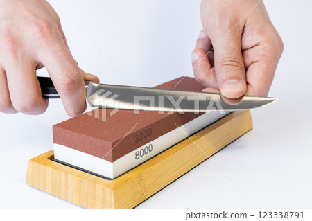 close up on white background on a table, man hands sharpen a steel knife protection against cuts, a red medium-hard stone for sharpening knives 123338791
