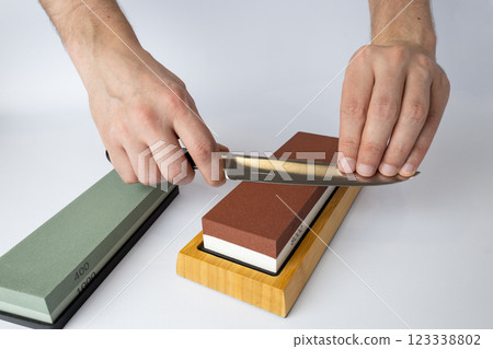 close up on white background on a table, man hands sharpen a steel knife protection against cuts, a red medium-hard stone for sharpening knives a green whetstone lies nearby close up on white background on a table, man hands sharpen a steel knife protection against cuts, a red medium-hard stone for sharpening knives a green whetstone lies nearby 123338802