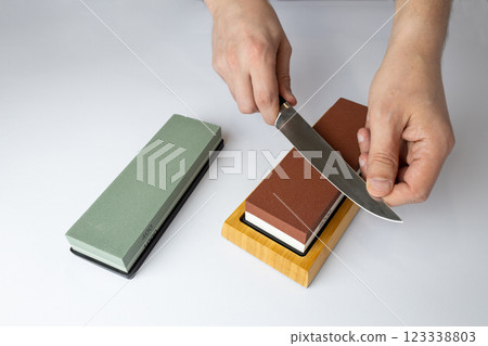 close up on white background on a table, man hands sharpen a steel knife protection against cuts, a red medium-hard stone for sharpening knives a green whetstone lies nearby 123338803
