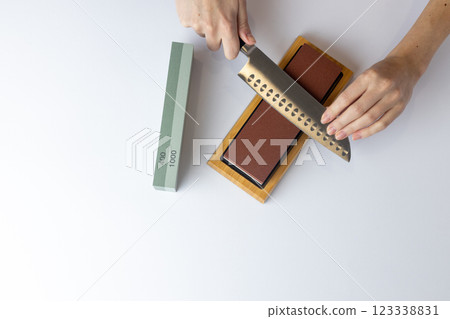 top shot on white background on a table, women's hands sharpen a steel knife, a lock under the arm, protection against cuts, a red medium-hard stone for sharpening knives green whetstone nearby 123338831