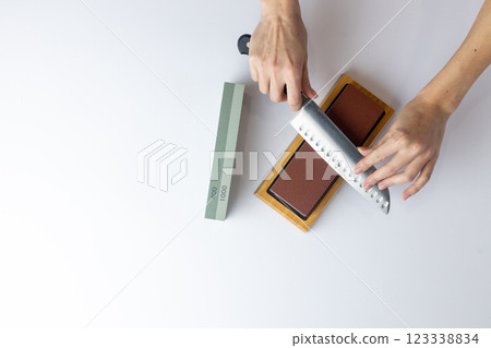 top shot on white background on a table, women's hands sharpen a steel knife, a lock under the arm, protection against cuts, a red medium-hard stone for sharpening knives green whetstone nearby 123338834