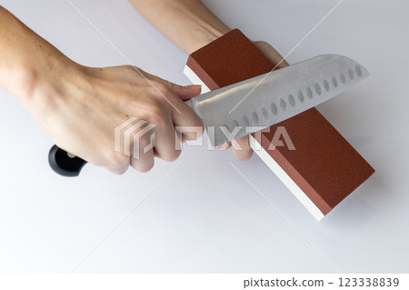 closeup on white background on a table, women's hands sharpen a steel knife, a lock under the arm, protection against cuts, a red medium-hard stone for sharpening knives 123338839
