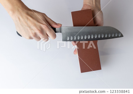 closeup on white background on a table, women's hands sharpen a steel knife, a lock under the arm, protection against cuts, a red medium-hard stone for sharpening knives closeup on white background on a table, women's hands sharpen a steel knife, a lock under the arm, protection against cuts, a red medium-hard stone for sharpening knives 123338841