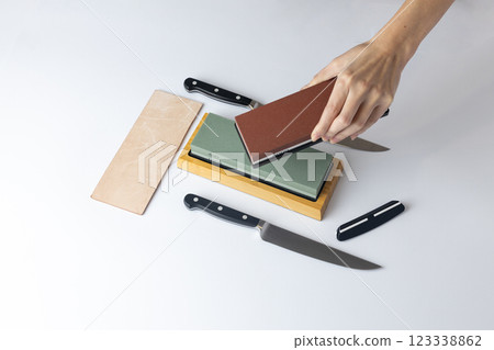 on white background on the counter lies the entire set of equipment for sharpening and polishing kitchen knives, the girl s hands hold a red stone of medium hardness, knives on the table 123338862