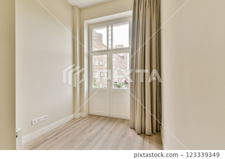 A bright, minimalist interior room featuring a large window with sheer curtains. The wooden floor complements the light-colored walls, creating an inviting atmosphere. A bright, minimalist interior room featuring a large window with sheer curtains. The wooden floor complements the light-colored walls, creating an inviting atmosphere. 123339349