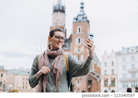 Beautiful stylish woman walking on Market Square in Krakow on autumn day and holding mobile phone. Phone Communication. Urban lifestyle concept. Check social networks, send sms. Beautiful stylish woman walking on Market Square in Krakow on autumn day and holding mobile phone. Phone Communication. Urban lifestyle concept. Check social networks, send sms. 123340597