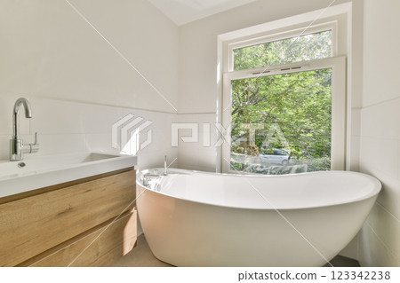 A sleek modern bathroom featuring a stylish freestanding bathtub, minimalist design elements, and large windows allowing natural light and a view of greenery outside. 123342238