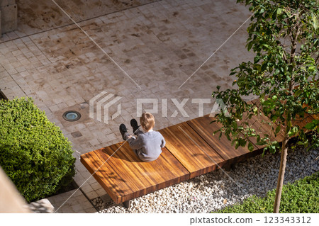 The image features an overhead view of an outdoor setting with a person sitting on a wooden bench. The person appears to be wearing light-colored clothes. The bench is surrounded by a neatly 123343312