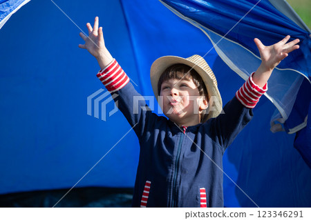 A young boy wearing a blue jacket and a straw hat is standing in front of a blue tent A young boy wearing a blue jacket and a straw hat is standing in front of a blue tent 123346291
