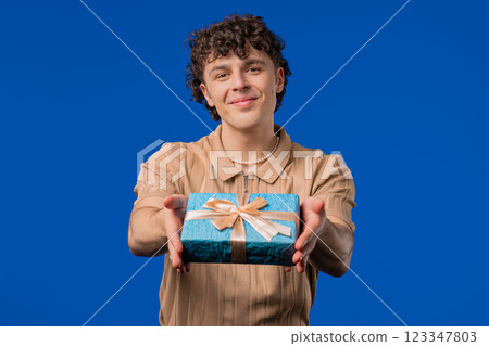 Man gives gift box by hands to camera on blue background. Guy smiling Man gives gift box by hands to camera on blue background. Guy smiling 123347803