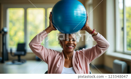 Poster with an elderly woman smiling while exercising and lifting a blue fitness ball over her head, supportive therapy providing safe and active exercise for older adults Poster with an elderly woman smiling while exercising and lifting a blue fitness ball over her head, supportive therapy providing safe and active exercise for older adults 123348047