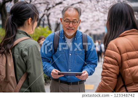 older man with a tablet in his hands talks to young women on an alley with blooming sakura trees, a man clarifies the route of a walk in the park, the concept of an active life of pensioners older man with a tablet in his hands talks to young women on an alley with blooming sakura trees, a man clarifies the route of a walk in the park, the concept of an active life of pensioners 123348742