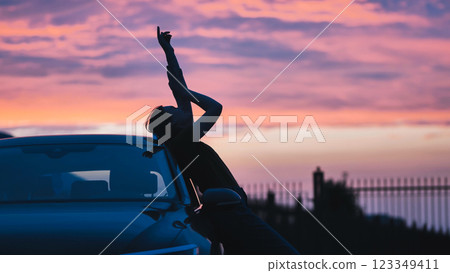 Elegant silhouette leaning against vehicle, arm raised, capturing dramatic sunset hues with feminine confidence 123349411