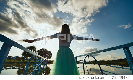 Young woman standing on bridge, arms spread wide, absorbing golden sunset light with peaceful expression 123349484