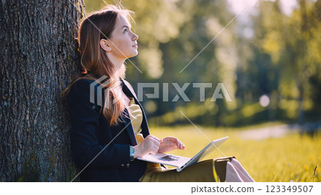 Young businesswoman relaxing under a tree in a park, using her laptop on a sunny day, enjoying the blend of nature and productivity 123349507