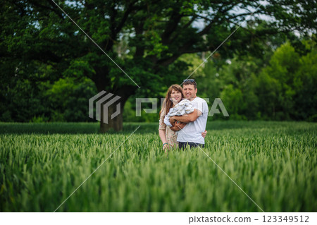 Young family standing barefoot in lush wheat field, cradling newborn near spreading tree under golden summer sunlight Young family standing barefoot in lush wheat field, cradling newborn near spreading tree under golden summer sunlight 123349512