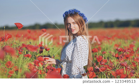 Blonde woman in white polka dot dress, blue floral headpiece, caressing red poppies under bright summer sunlight Blonde woman in white polka dot dress, blue floral headpiece, caressing red poppies under bright summer sunlight 123349513