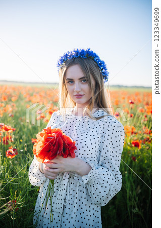 Young woman wearing cornflower wreath, holding bright poppy bouquet, standing amid flowering poppy landscape Young woman wearing cornflower wreath, holding bright poppy bouquet, standing amid flowering poppy landscape 123349699