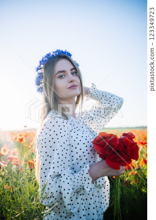 Blonde woman wearing cornflower wreath, holding poppy bouquet while standing in sunlit summer meadow Blonde woman wearing cornflower wreath, holding poppy bouquet while standing in sunlit summer meadow 123349723