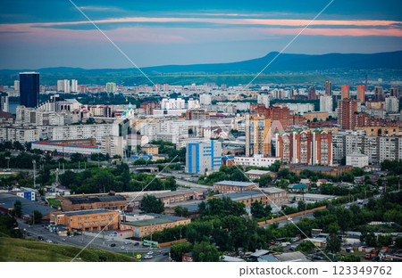 Krasnoyarsk cityscape revealing urban landscape, residential zones, winding streets, verdant greenery, mountain silhouettes against dramatic sunset lighting 123349762