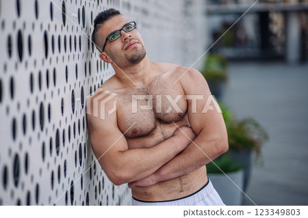 Shirtless athlete with glasses and crossed arms leaning against modern metallic wall, resting after exercising outdoors in russia 123349803