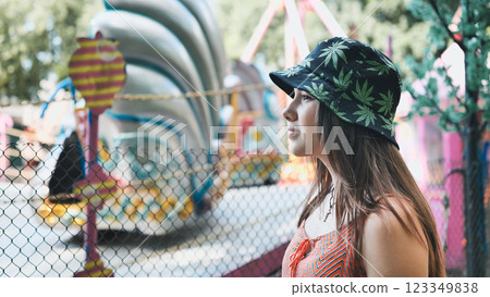 Girl wearing a bucket hat with cannabis leaf prints looking at an amusement park ride in summer 123349838