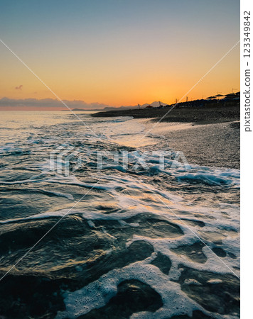 Soft waves lapping rocky shoreline during vibrant sunset along Cretan coastline, capturing serene Mediterranean summer evening atmosphere Soft waves lapping rocky shoreline during vibrant sunset along Cretan coastline, capturing serene Mediterranean summer evening atmosphere 123349842