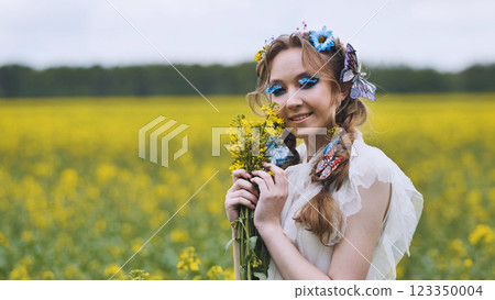 Model wearing white dress and butterfly decorations holding rapeseed bouquet in a yellow field during a cloudy day 123350004