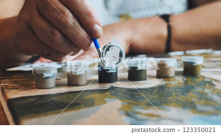 Artist's hand hovering near paint pot, grasping paintbrush, preparing to fill numbered canvas section with vibrant color during creative hobby activity 123350021