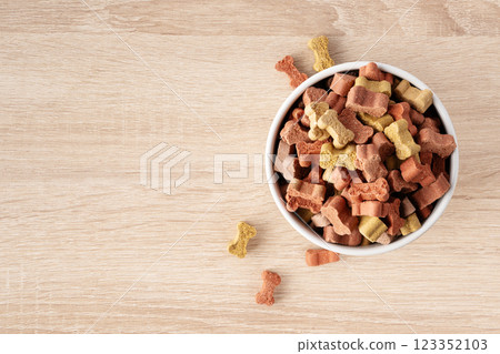 Dry dog food in a shape of bones in bowl on wooden floor background top view with copy space. Plate full of mineral healthy dog food. Pet snack in white bowl on floor. 123352103