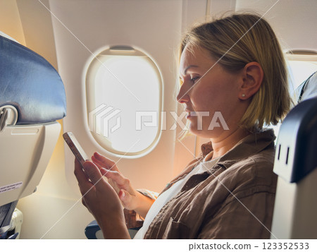 Passenger woman is flying in plane. Girl using phone sitting by airplane window. Traveling female with smartphone inside plane enjoying flight. Woman using wifi connection on board. Traveling girl 123352533