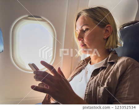 Passenger woman is flying in plane. Girl using phone sitting by airplane window. Traveling female with smartphone inside plane enjoying flight. Woman using wifi connection on board. Traveling girl 123352534
