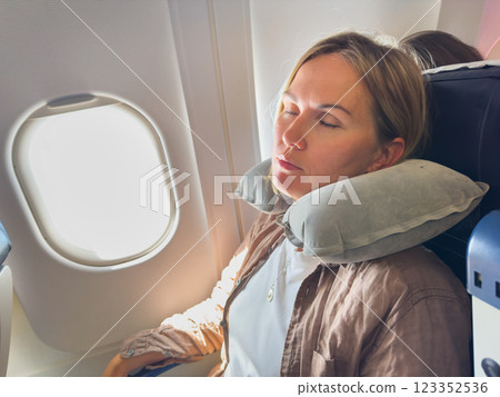 Rest on plane. Young woman napping sitting by porthole in business jet using neck pillow for travel. Passenger girl flying in plane. Girl sleeping in airplane going on trip vacation. Traveling girl 123352536