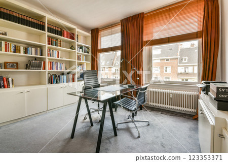A stylish office space featuring a glass desk, ergonomic chairs, and extensive bookshelves. Natural light floods through large windows, enhancing the workspace ambiance. A stylish office space featuring a glass desk, ergonomic chairs, and extensive bookshelves. Natural light floods through large windows, enhancing the workspace ambiance. 123353371