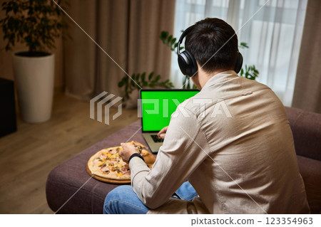 Young man sitting on sofa and eating pizza while using laptop at home 123354963