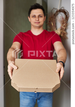 smiling delivery man in red t-shirt giving food order, holding pizza box standing at doorway smiling delivery man in red t-shirt giving food order, holding pizza box standing at doorway 123354975