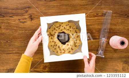 Flat lay. Carefully placing the gingerbread bundt cake, adorned with salted caramel frosting and gingerbread sprinkles, into a white paper bundt cake box with a clear window for gifting. 123355112