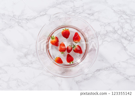 Ripe strawberries are submerged in water within a large glass mixing bowl, a step in washing the fruit to ensure cleanliness and longevity before storage or consumption. 123355142